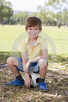 Kid 7 or 8 years old enjoying happy playing football soccer at grass city park field posing smiling proud sitting on the ball in