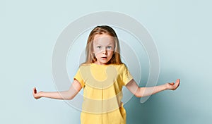 Kid in yellow t-shirt. She spreads her hands wide apart like holding something, looking surprised, posing on turquoise background