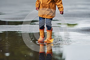 Kid in yellow waterproof cloak and boots playing with paper handmade boat toy outdoors after the rain