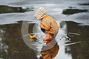 Kid in yellow waterproof cloak and boots playing with paper handmade boat toy outdoors after the rain