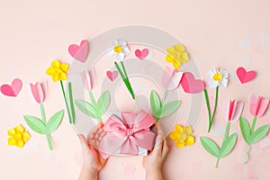 Kids hands holding gift present box with pink bow decorated paper hearts and tulip flowers on pink pastel table top view. Flat lay