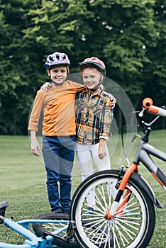 Kids in helmets embracing and looking at camera while standing near bicycles at park