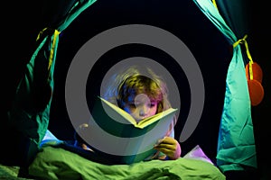 Kids in kids tent reading a book in the dark home. Child boy reading a book lying on the bed. Dreaming child read