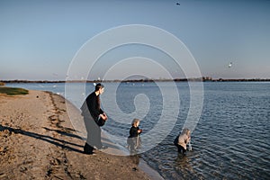 Kids and mom running in water on river bank in autumn. Boy is drinking river water. Disobedience. Happy family on the