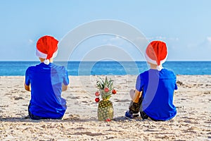 Kids in red Santa hats sitting at sunny beach during Christmas vacation and looking to sea