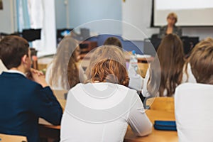 Kids in school writing and taking notes, teens pupils behind desks during the lesson listen to teacher lecture, classroom with