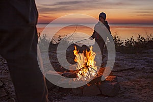 Kids sit near fire on autumn seashore after sunset, warm their hands, have fun together, outside
