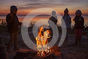 Kids sit near fire on autumn seashore after sunset, warm their hands, have fun together, outside