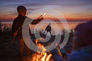 Kids sit near fire on autumn seashore after sunset, warm their hands, have fun together, outside