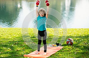 Kids sport. Cute little boy doing exercises with dumbbells in green park. Portrait of sporty child with dumbbells. Happy