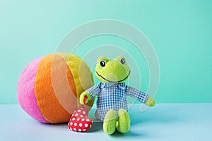 Kids Toys Small Plush Frog Holding Red Heart Multicolored Textile Soft Ball on Blue Green Background. Banner Placeholder Charity