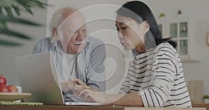 Kind Asian caregiver teaching senior man using computer typing and talking in kitchen in house