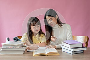 A kind Asian female teacher helps a young, adorable girl read a book and do homework