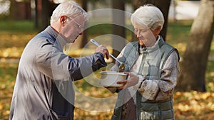 Kind caucasian retired grey-haired man helps people in need by sharing his homemade soup with elderly homeless woman.