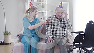 Kind Caucasian woman giving birthday cake to old grey-haired man in nursing home. Depressed retiree taking off party hat