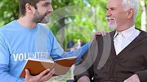 Kind male volunteer reading book to smiling old man in wheelchair, assistance