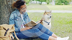Kind mixed race woman is reading book in park and stroking her dog sitting on lawn under tree together. Intelligent