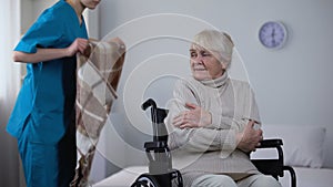 Kind nurse covering with blanket handicapped old woman shivering from cold