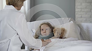 Kind pediatrician examining sick little girl at home, doctor comforting ill kid