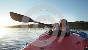 Kindergartener child learns to sail red kayak along large tranquil lake against forest at bright summer sunset