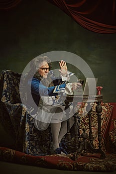 King, man in classical baroque-style attire drink wine while holding meeting online against vintage studio background.