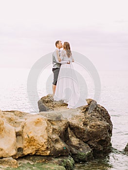 The kissing newlyweds standing on the cliff at the background of the sea.