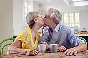 Kissing Retired Couple Sitting Around Table At Home Having Morning Coffee Together