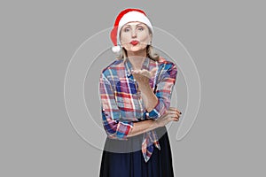 Kissing you! Portrait of kindly beautiful adult woman in red santa cap and checkered shirt standing and sending you air kiss,