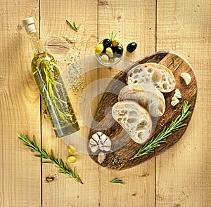 Kitchen chopping board with sliced fresh ciabatta, sprigs of rosemary, garlic, green and black olives. Bottle of olive