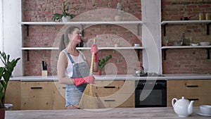 Kitchen cleaning, happy housewife in denim overalls and gloves dance and plays on broom like guitar