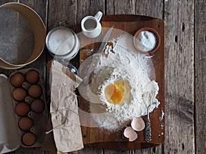 On a kitchen cutting board, a broken egg in flour with ingredients for making dough. Different kitchen utensils