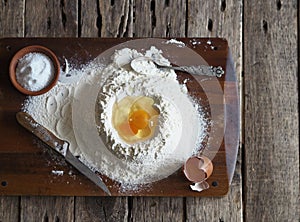 On a kitchen cutting board, a broken egg in flour with ingredients for making dough. Different kitchen utensils