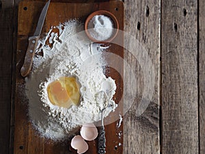 On a kitchen cutting board, a broken egg in flour with ingredients for making dough. Different kitchen utensils