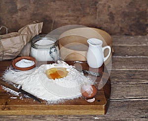 On a kitchen cutting board, a broken egg in flour with ingredients for making dough. Different kitchen utensils