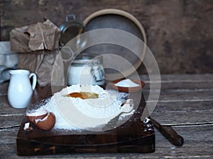 On a kitchen cutting board, a broken egg in flour with ingredients for making dough. Different kitchen utensils