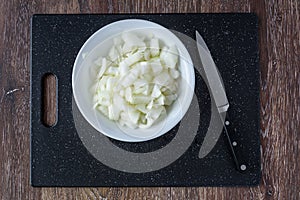 Kitchen knife and a diced raw sweet onion on a black cutting board with white bowl