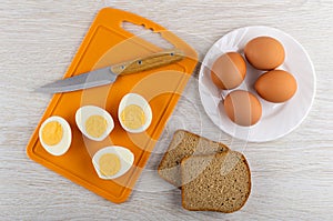 Knife, halves of peeled eggs on cutting board, pieces of bread, eggs in plate on wooden table. Top view