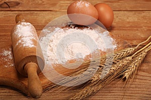 Kitchen rolling pin, wooden board with flour on wooden background, ears and eggs for making bread on table