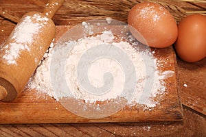 Kitchen rolling pin, wooden board with flour on wooden background, ears and eggs for making bread on table
