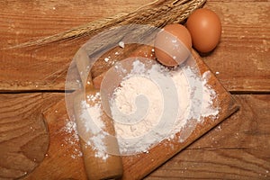 Kitchen rolling pin, wooden board with flour on wooden background, ears and eggs for making bread on table