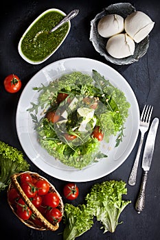 Kitchen table with fresh mozzarella salad, lettuce and arugula leaves, pesto sauce, fresh cherry tomatoes in a basket, flatlay