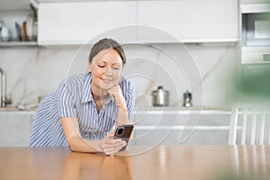 In the kitchen, teen woman leans on table with phone in hand, propping up chin with fist.