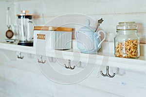 Kitchen wooden shelf with tea leaves in gold box and accessories, blue sugar bowl with spoon, strainer, press. Cup, mug hooks.