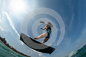 Kitesurfing girl in black sexy swimsuit with kite in sky on board in blue sea riding waves with water splash. Recreational