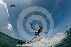 Kitesurfing girl in black sexy swimsuit with kite in sky on board in blue sea riding waves with water splash.