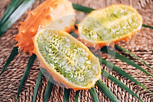 Kiwano or african horned melon with palm leaves on rattan background. Cutted hedged gourd, african horned cucumber, english tomato
