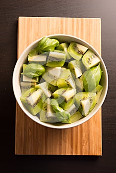 Kiwi fruit sliced segments with basil leaves in a round bowl on the wooden cutting board