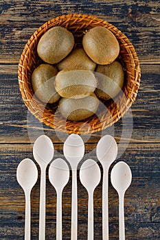 Kiwi fruits with spoons in a wicker basket on wooden background, top view