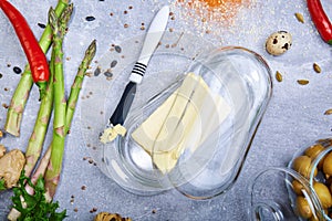 Knife and briquette of margarine. Butter in a glass box and vegetables on a table background. Healthy meal preparation.