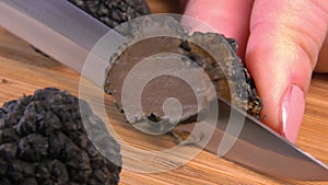 A knife cuts a slice from a black truffle on a wooden board